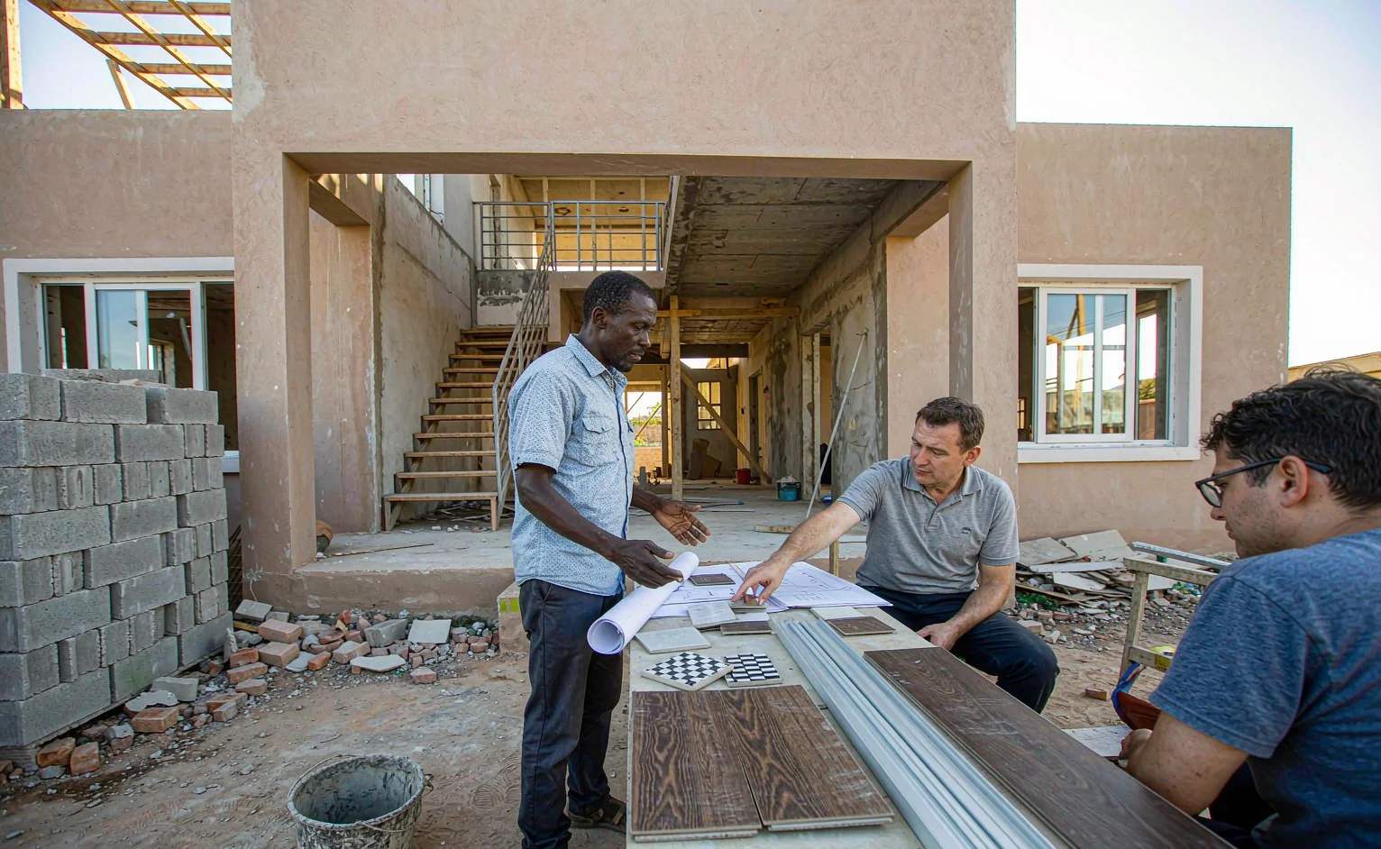 Engineer and property owner reviewing tile, aluminum profile and flooring samples on a table at a villa construction site in East Africa