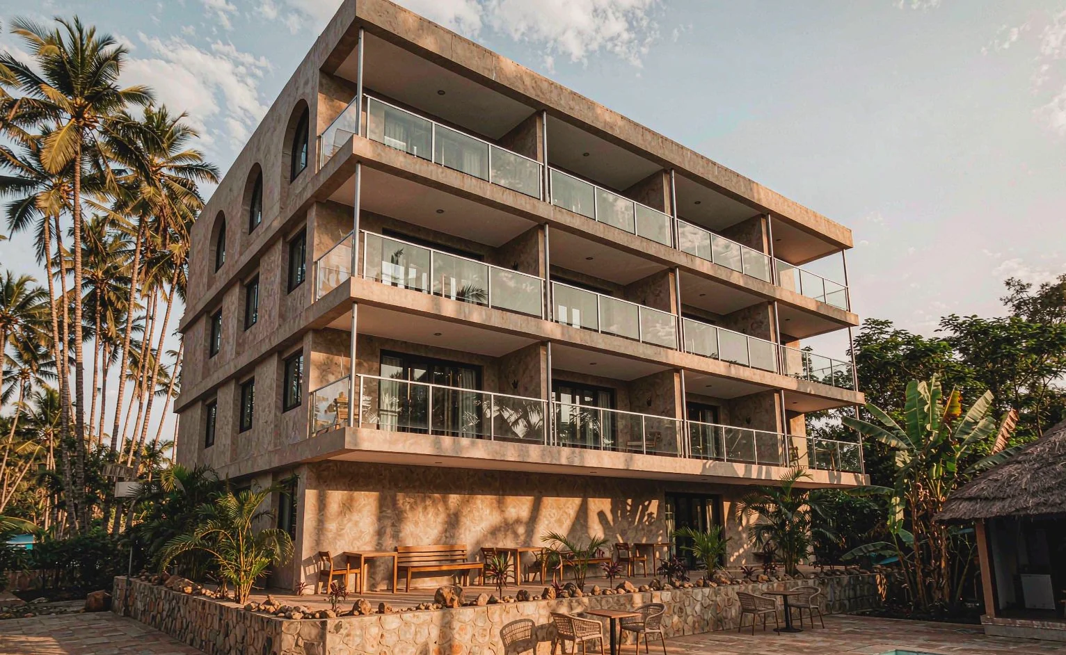 Four-storey boutique hotel exterior with glass balustrades, stone-look coating and tropical landscaping under strong sun