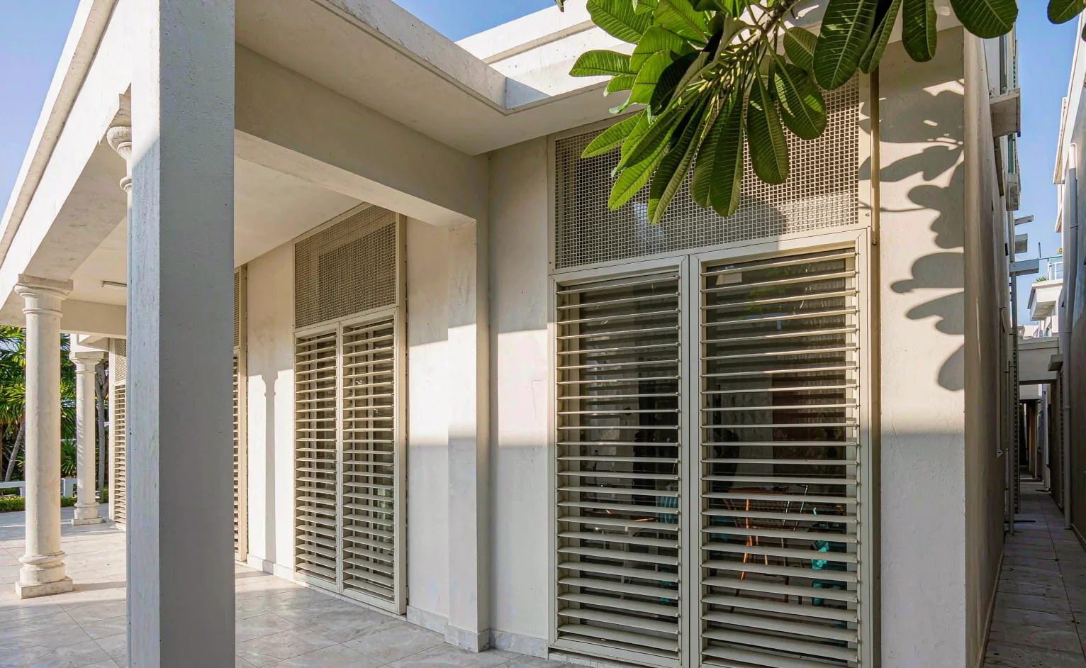 Tropical hotel facade in strong midday sun with external aluminium louvre shutters half closed over tall glazing under a deep overhang