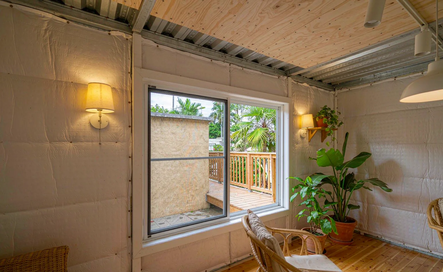 Interior of a converted shipping container home with insulated wall panels, plywood ceiling, large window and rattan furniture