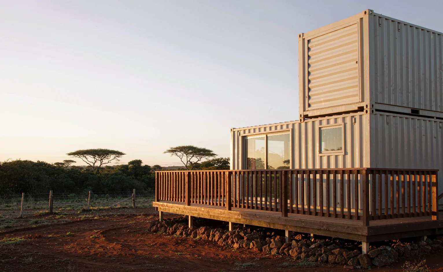 Two stacked converted shipping containers forming a home with wooden deck and full-height glass openings on red East African soil at golden hour