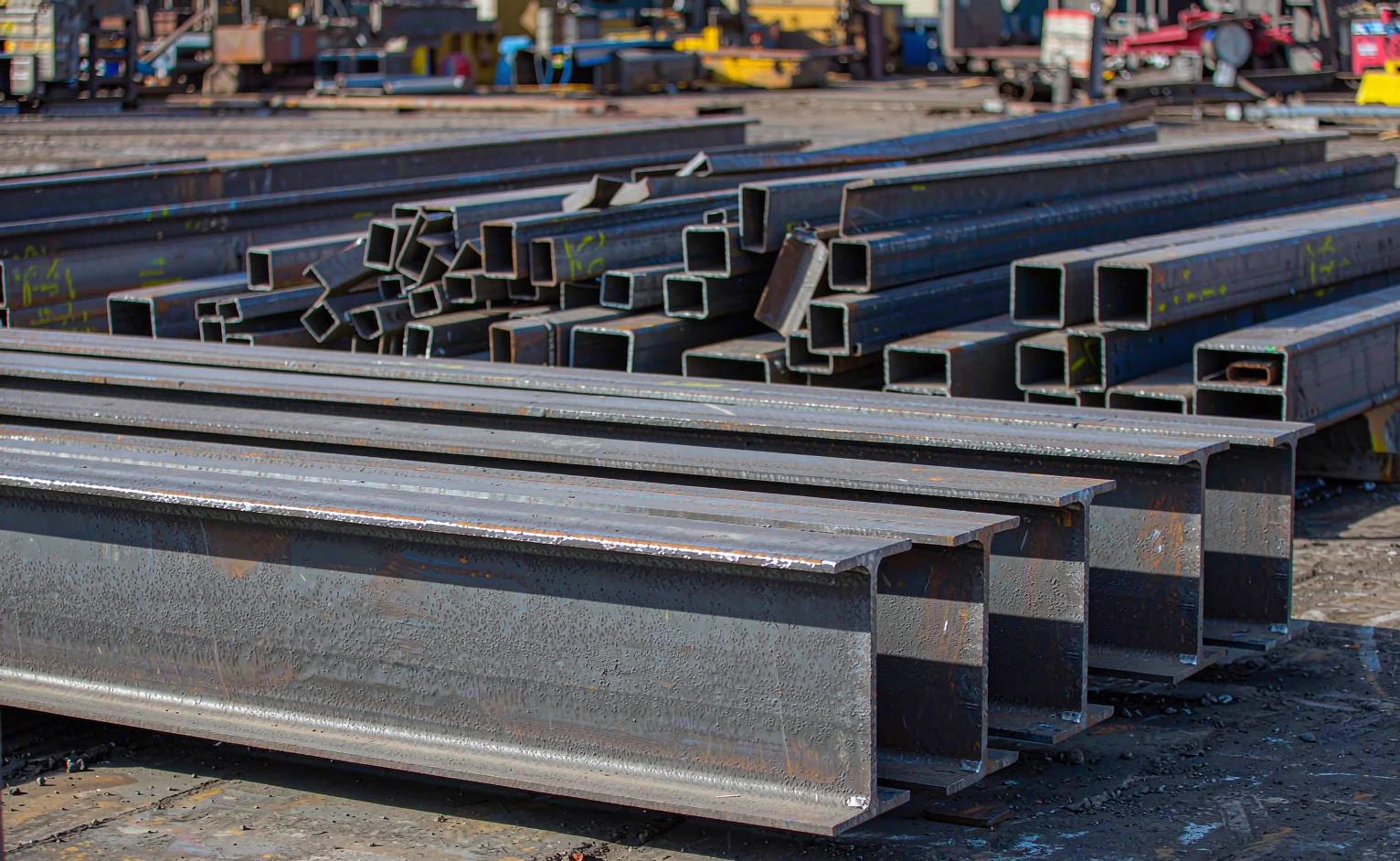 Stacked hot-rolled I-beams H-columns and square steel tubes in a fabrication yard awaiting fabrication into a portal frame
