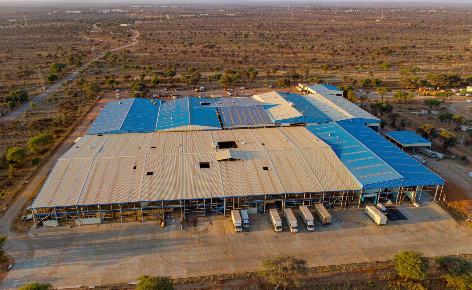 Aerial drone view of a completed steel warehouse complex with blue and off-white roof sheets and trucks on a concrete apron in East Africa